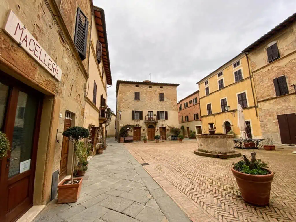 Empty Piazza Spagna in Pienza, Italy. Orange and yellow buildings frame the small piazza. There is a stone well in the middle. Sign for Macelleria on building on left.