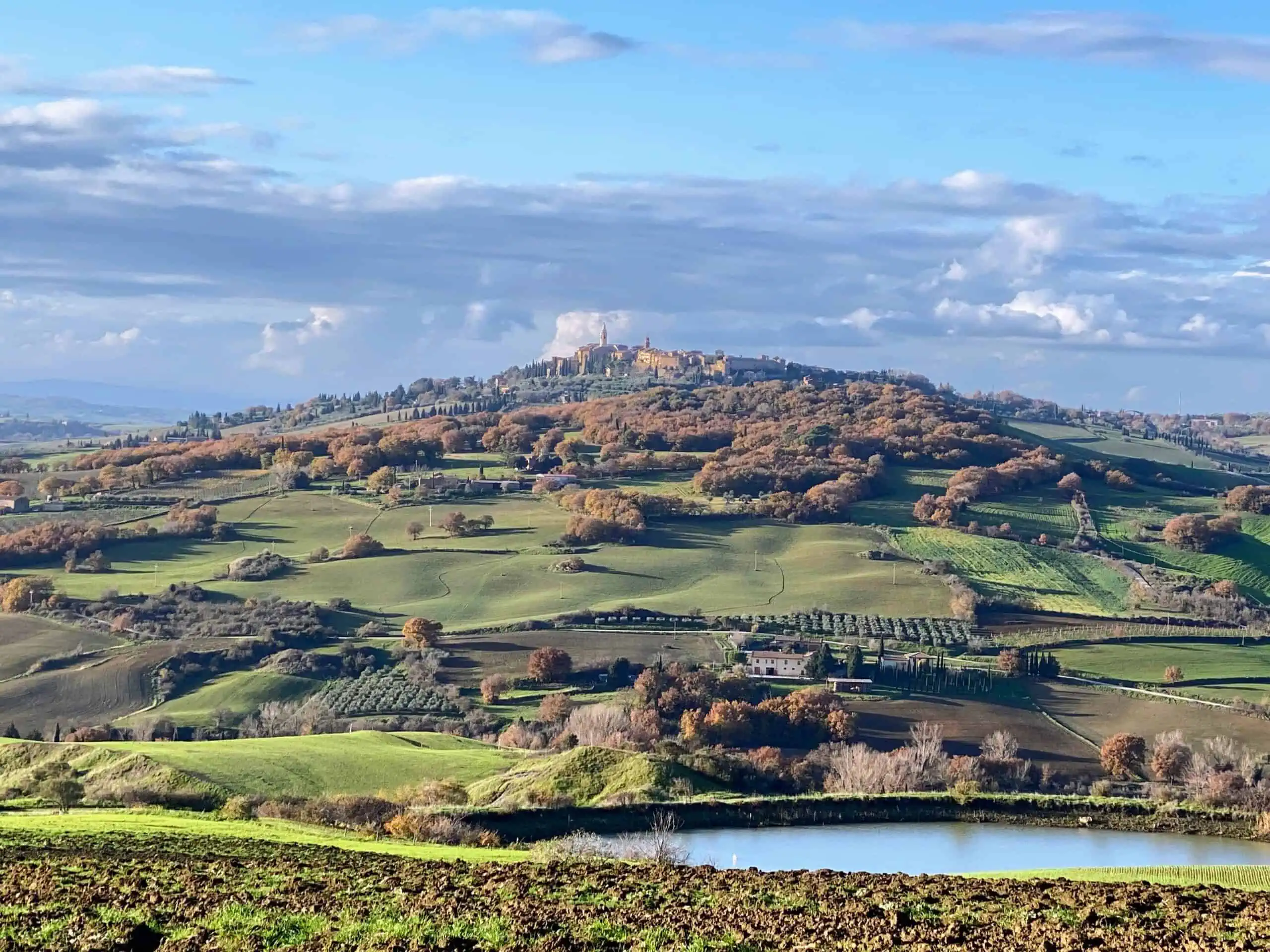 View of Pienza, Italy from a distance. You can see the small hilltop town in the distance. It's surrounded by woods and green rolling hills. Small pond in the foreground.