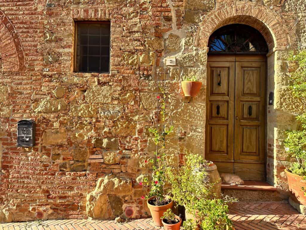 Beautiful early evening sun shines on old red brick wall and entrance to a home in Monticchiello, Italy. A cat naps on the doorstep and plants decorate the entrance.