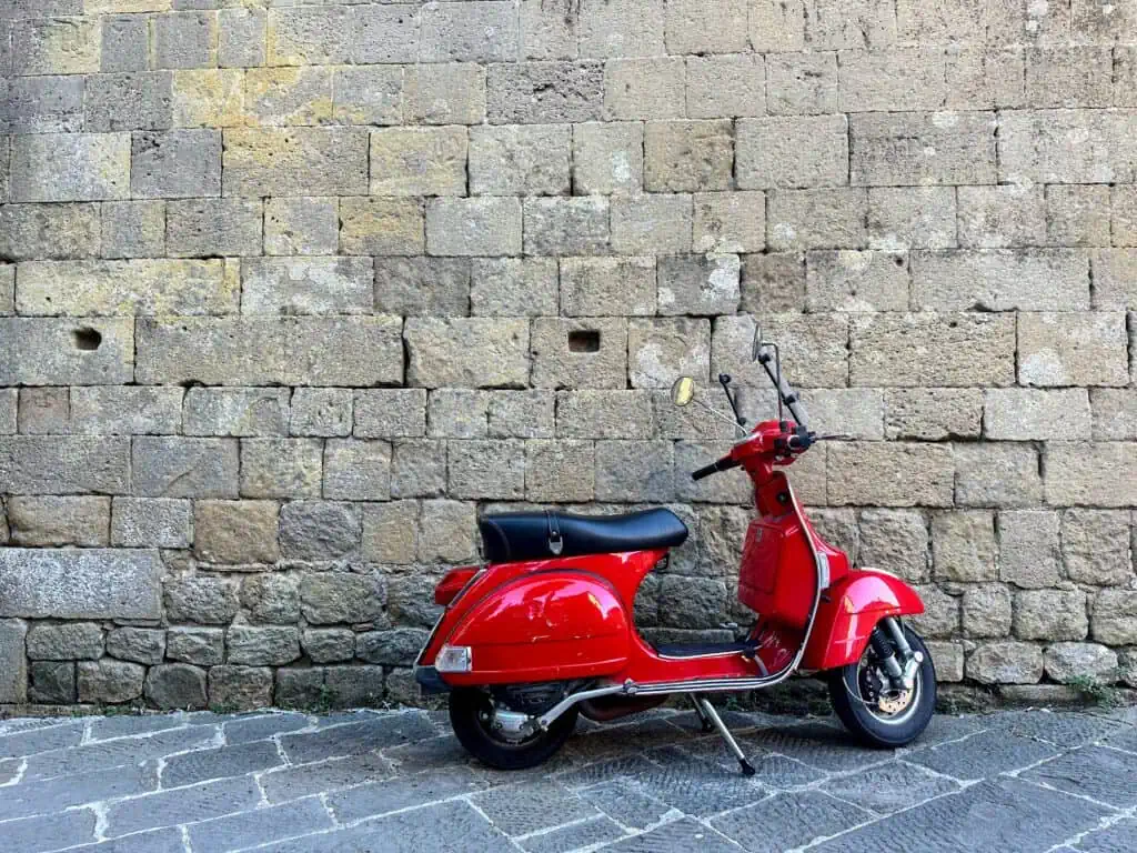 Bright red vespa sits in front of a stone wall in Tuscany.
