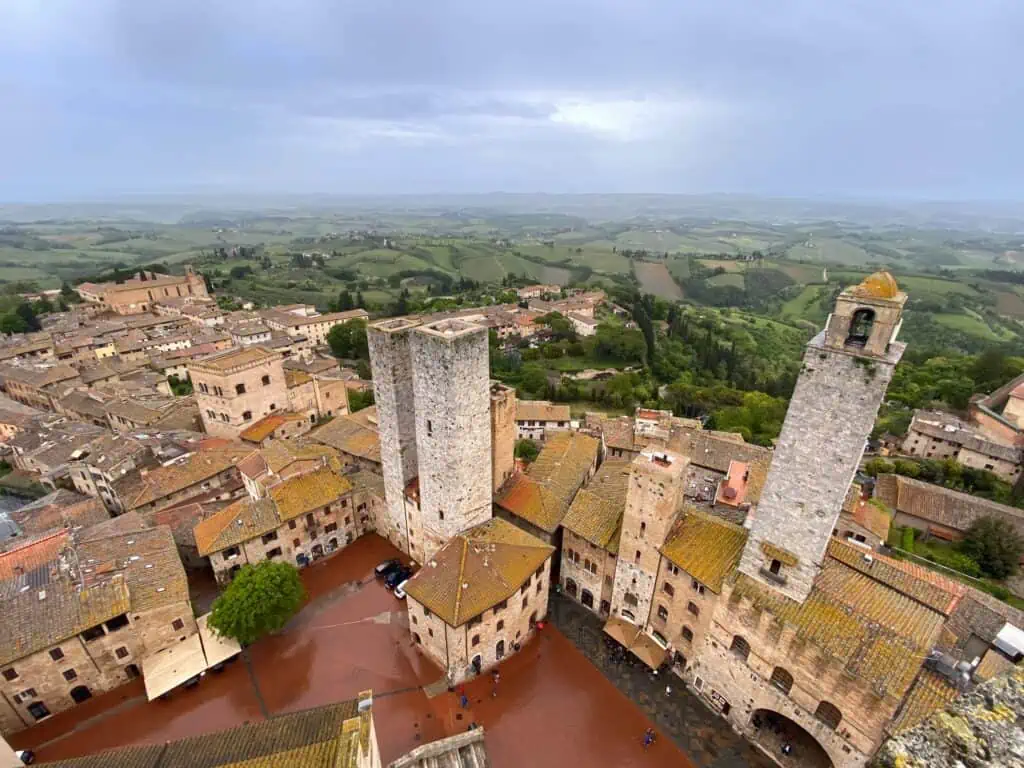 View of green Tuscan countryside on a stormy day. You can also see tall towers and a piazza and buildings in an Italian town.