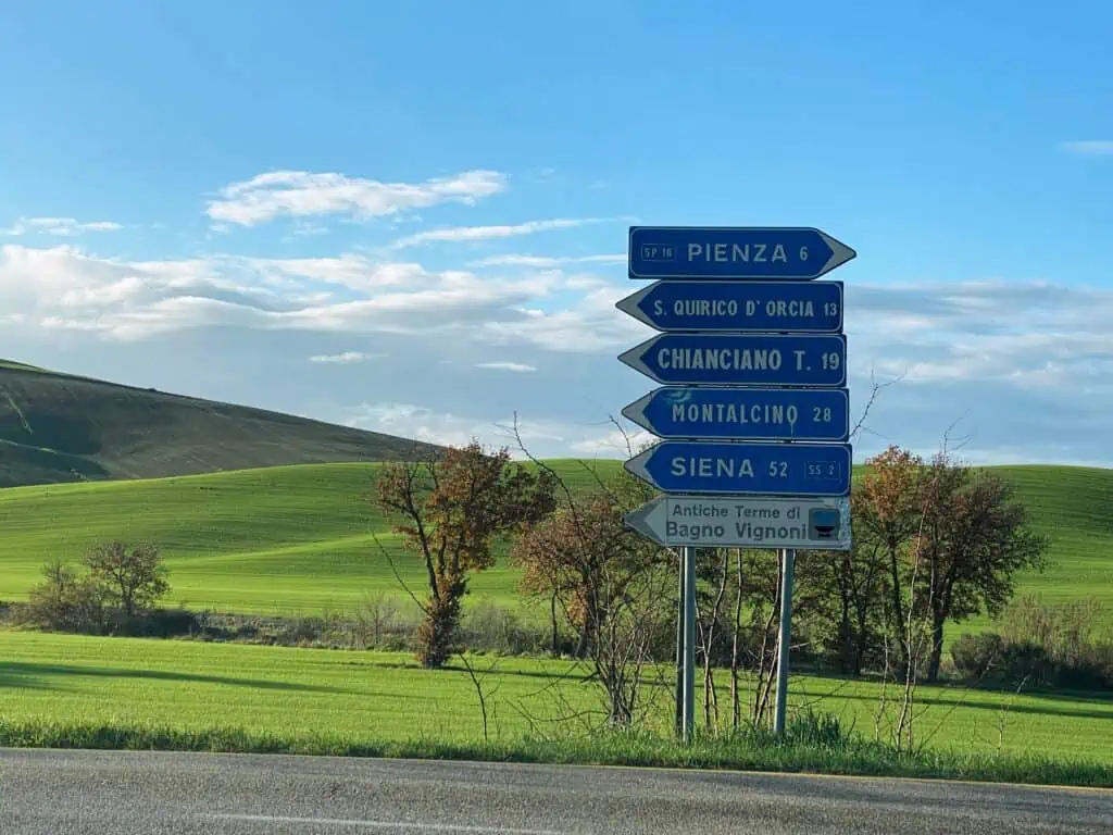 Blue road signs point to signs for village in the Val d'Orcia in Italy. In the background is bright green grass and blue sky and rolling hills.
