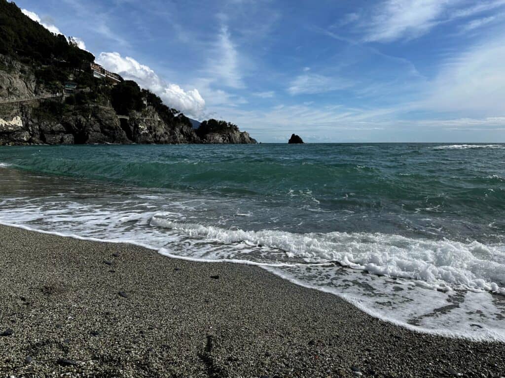 Waves crash on pebbly beach. Rocky cliffs in back right.