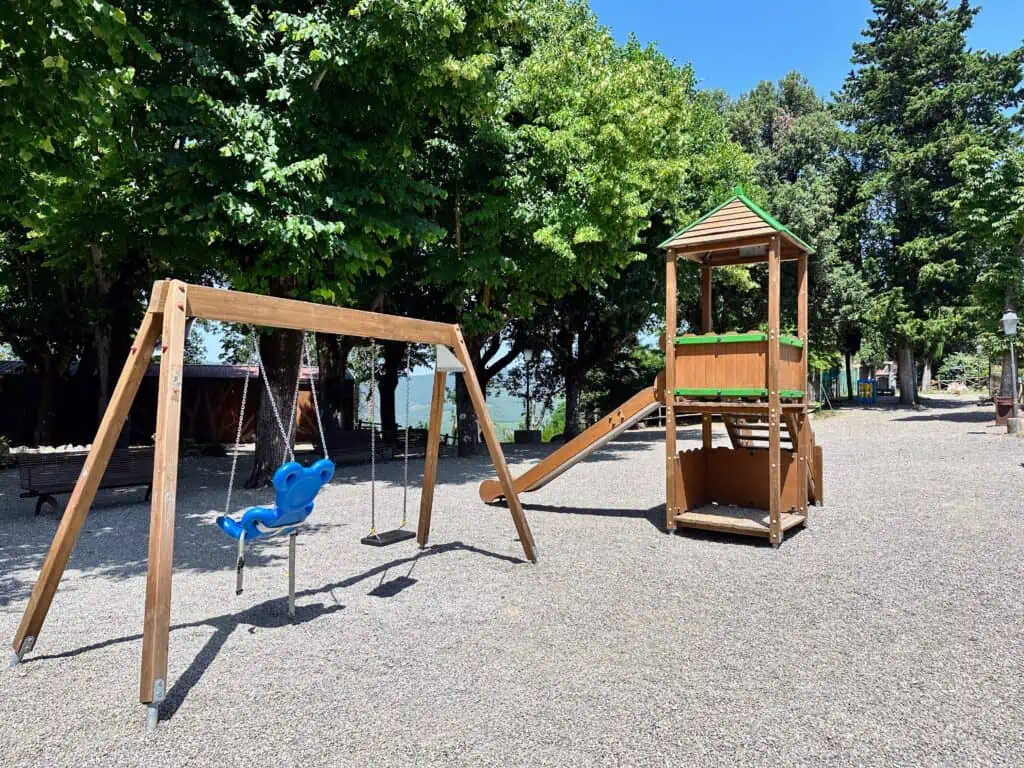 Wooden toy structures including slide and swings at a playground in Italy. Gravel underneath and trees provide some shade.