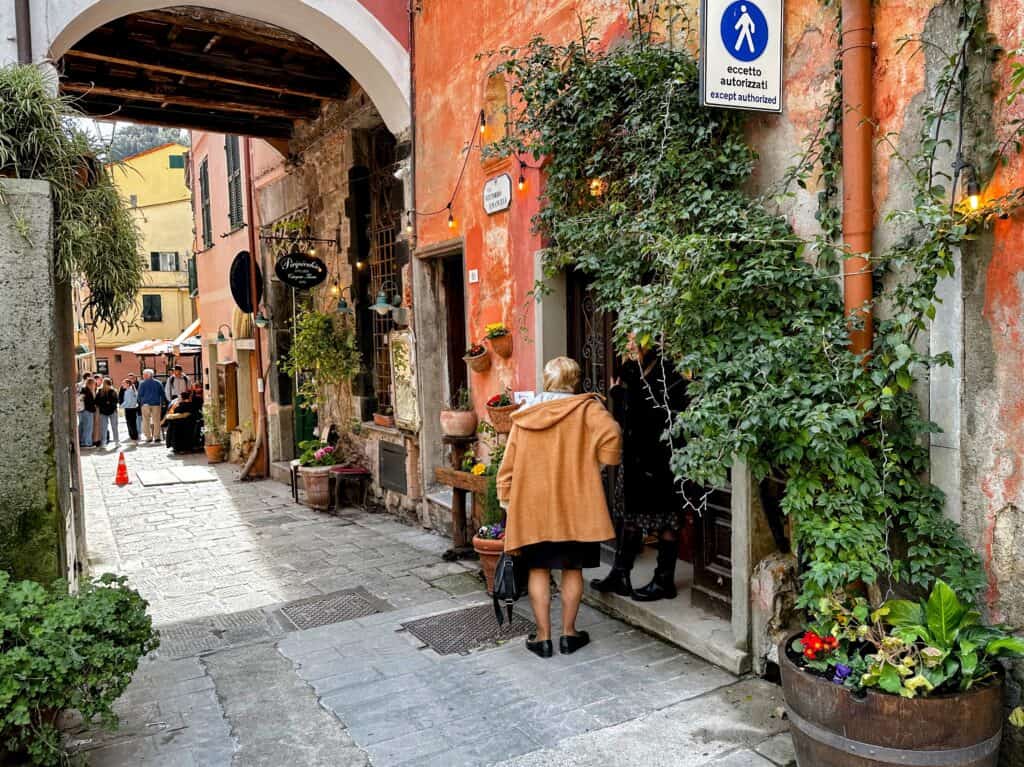Woman in orange coat stands next to orange building covered in vines in Monterosso al Mare's old town. Arch on left and people in street past arch.