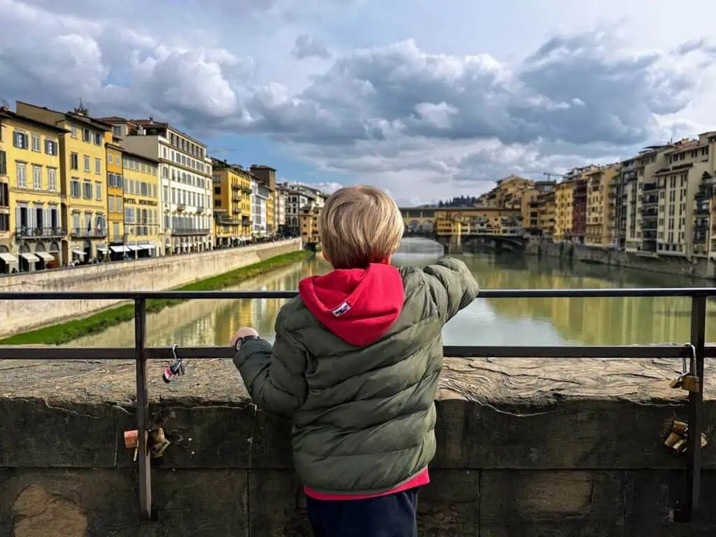 Boy wearing green coat on a bridge in Florence, looking out at the Arno River and the Ponte Vecchio on a cloudy day.