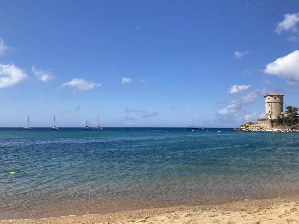 Campese beach, with sand, turquoise water, and sailboats in the water. Tower and small piece of land on right.