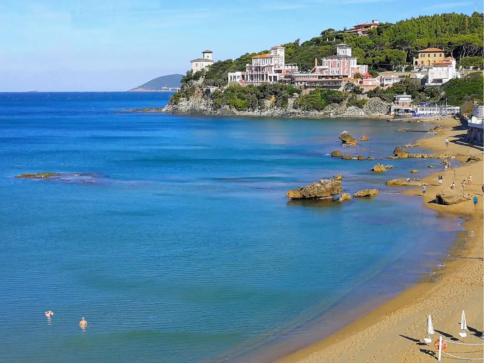 People swim and walk on the beach. Rocks in the water, buildings on a promontory in background.