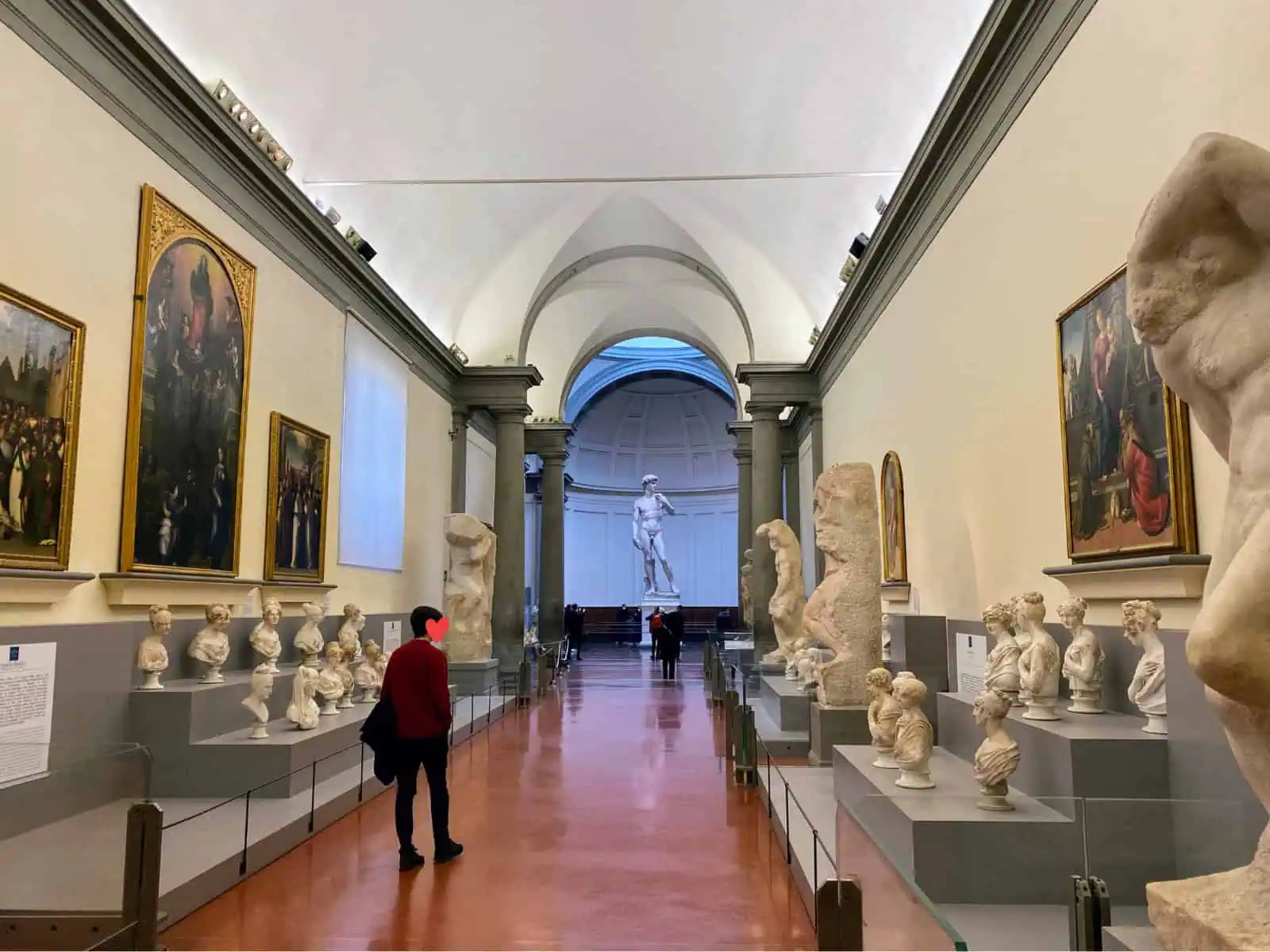 An empty Accademia Gallery in Florence, Italy. One woman walks down hallway filled with busts and sculptures toward Michelangelo's David statue at the end. A few others look at the David statue.