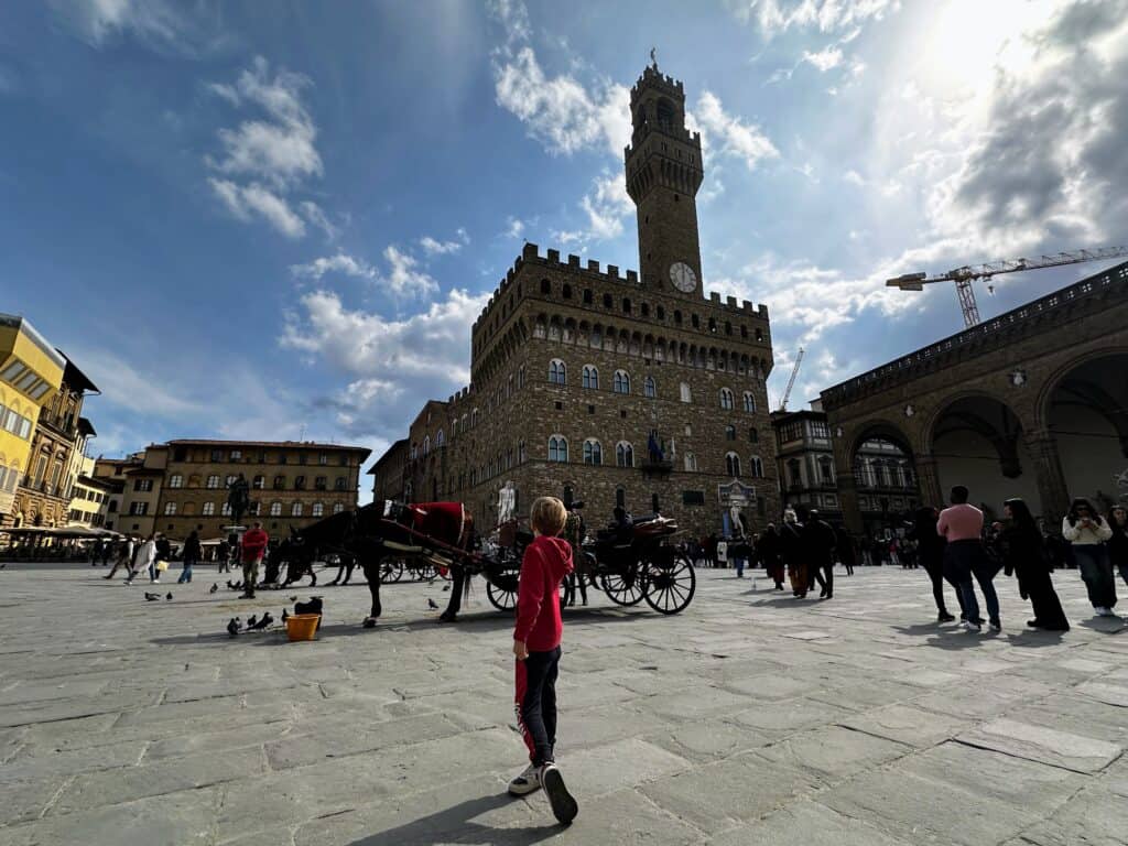 Boy in red stands in Piazza alla Signoria in Florence and looks toward Palazzo Vecchio. A few people are scattered around the piazza and there is a horse and carriage in the center of the piazza. It's a sunny day with a few white clouds.