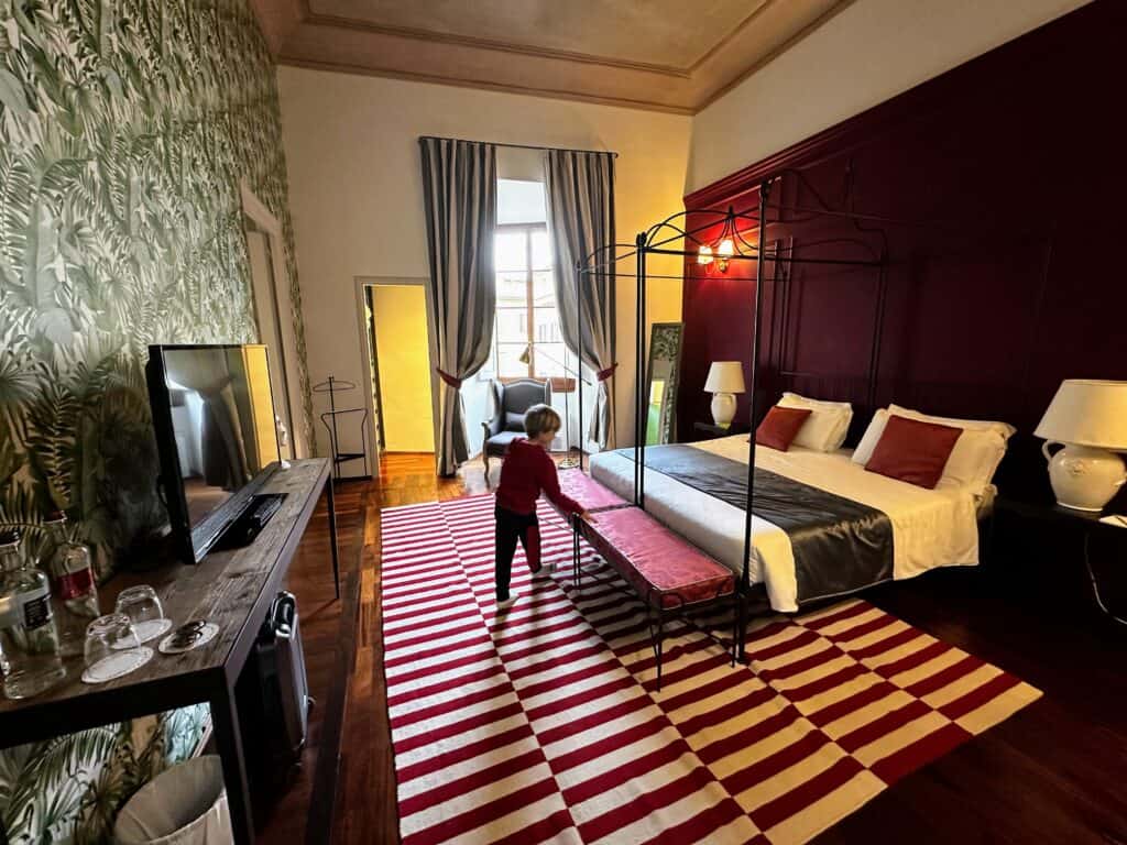 Boy looks at bed inside a room at Peruzzi Urban Residences in Florence, Italy. The wooden floor has a large red and white carpet. There is a window in the back and a tv and wooden table on the left.