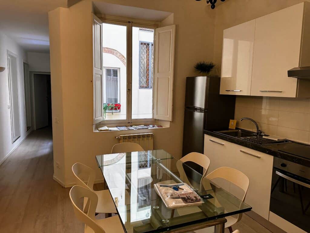 Kitchen with glass dining table and white chairs in an aparment in Florence, Italy. There is a window in the kitchen that looks out into an internal courtyard.