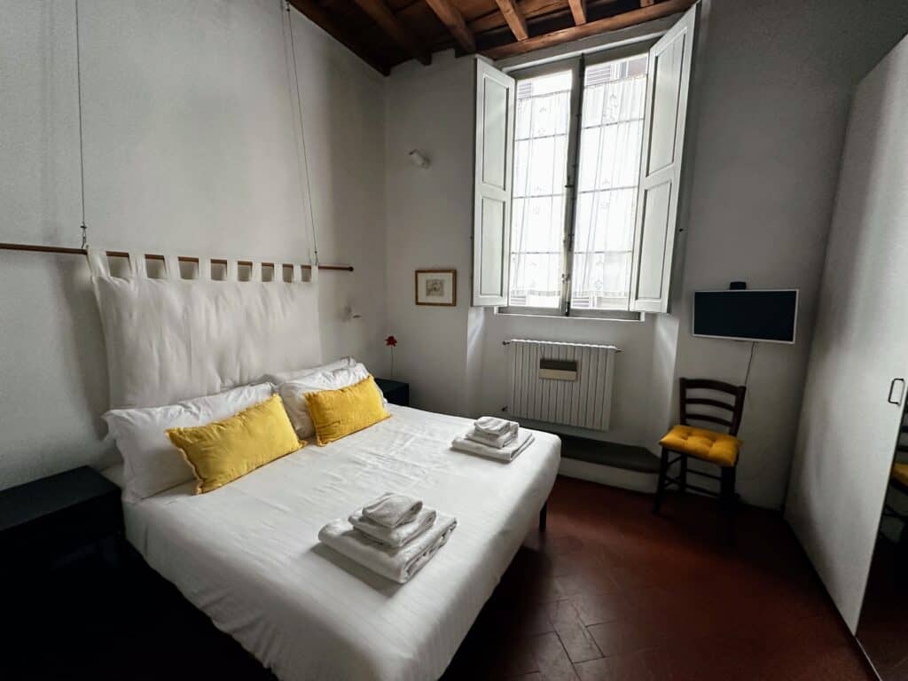 Bedroom with terracotta floor in Florence, Italy. White bedspread and pillows and two yellow pillows on bed. Window on far wall with lace shades. Wooden chair and small tv in right corner.