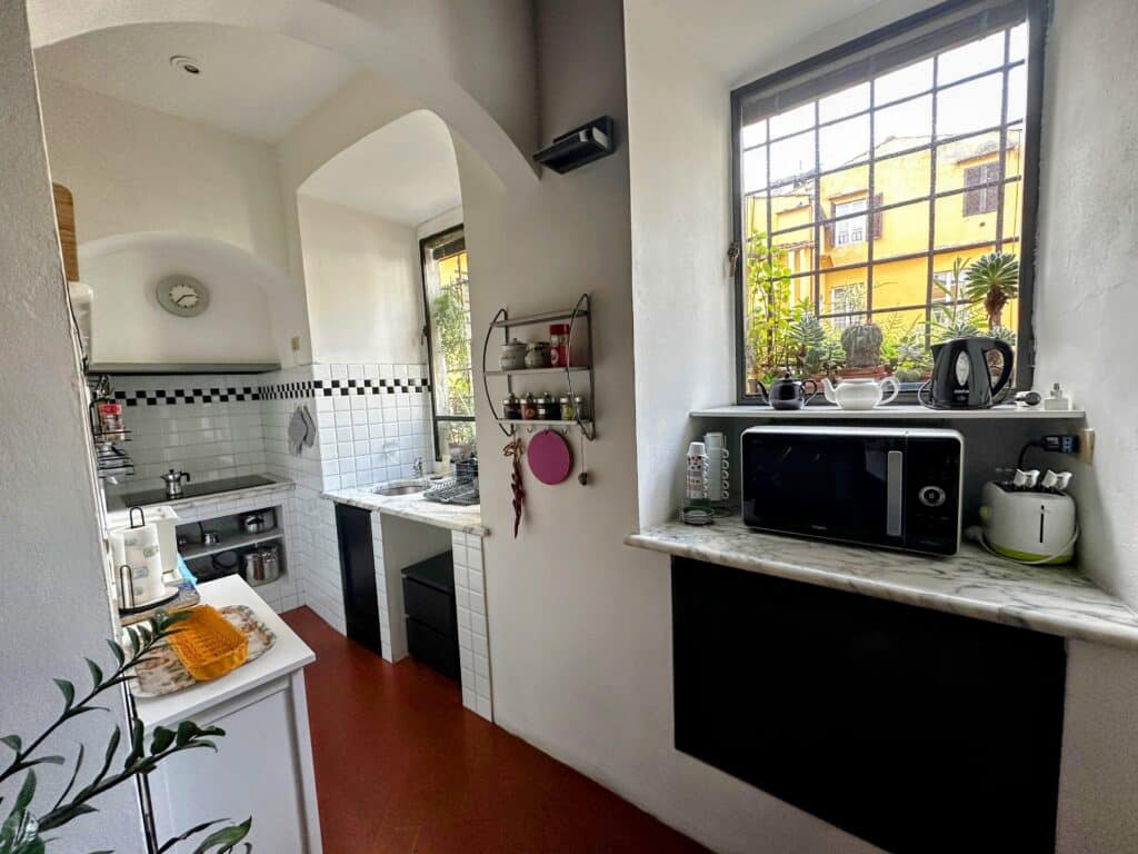 Kitchen in apartment in Florence, Italy. White tile backsplash, appliances like microwave and sink and windows looking out onto other yellow buildings. Plants on the windowsills.