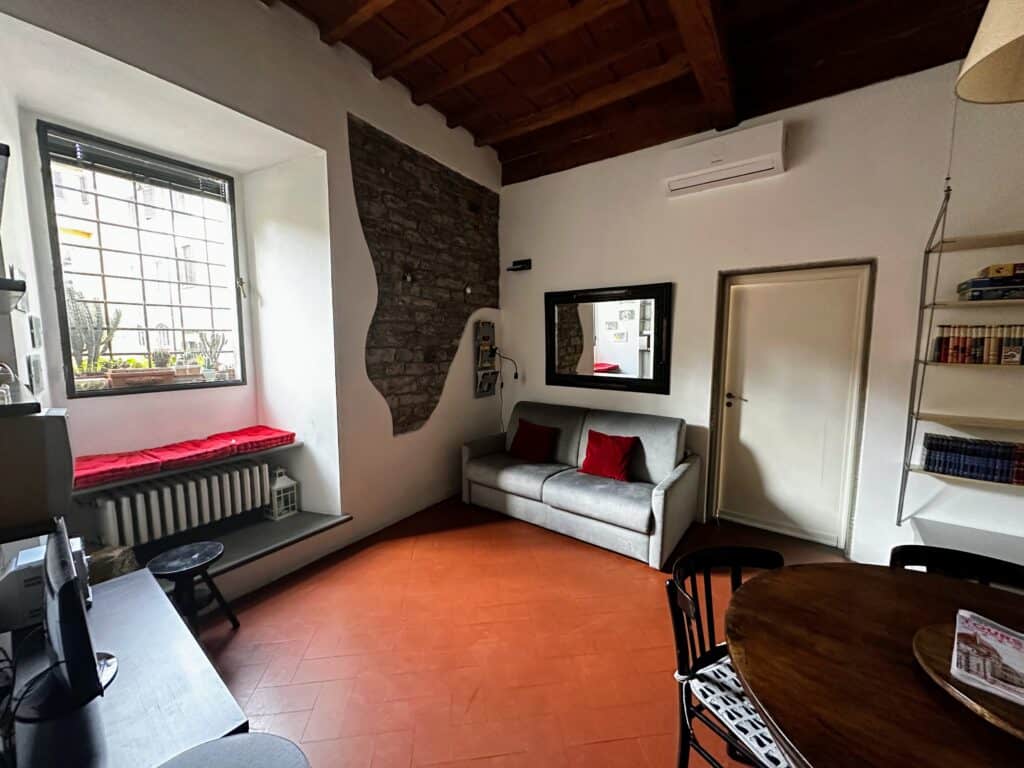 Terracotta floor in a living room in an apartment in Florence, Italy. Grey couch on back wall and part of wall on left is exposed brick. Window on left looks at another building. Window seat with red cushions below the window. Wooden table and chairs on right.