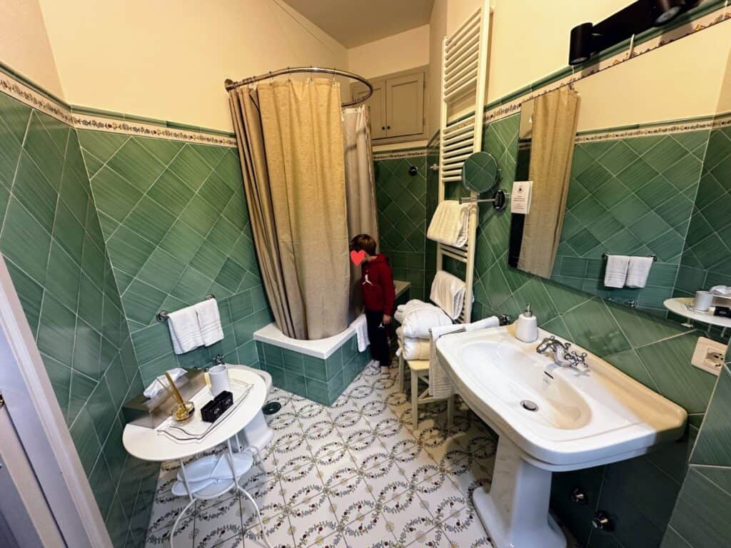 Boy looks at bathtub in green tiled bathroom at Peruzzi Residences in Florence, Italy. Bathroom also has sink and mirror and bidet and small white table.