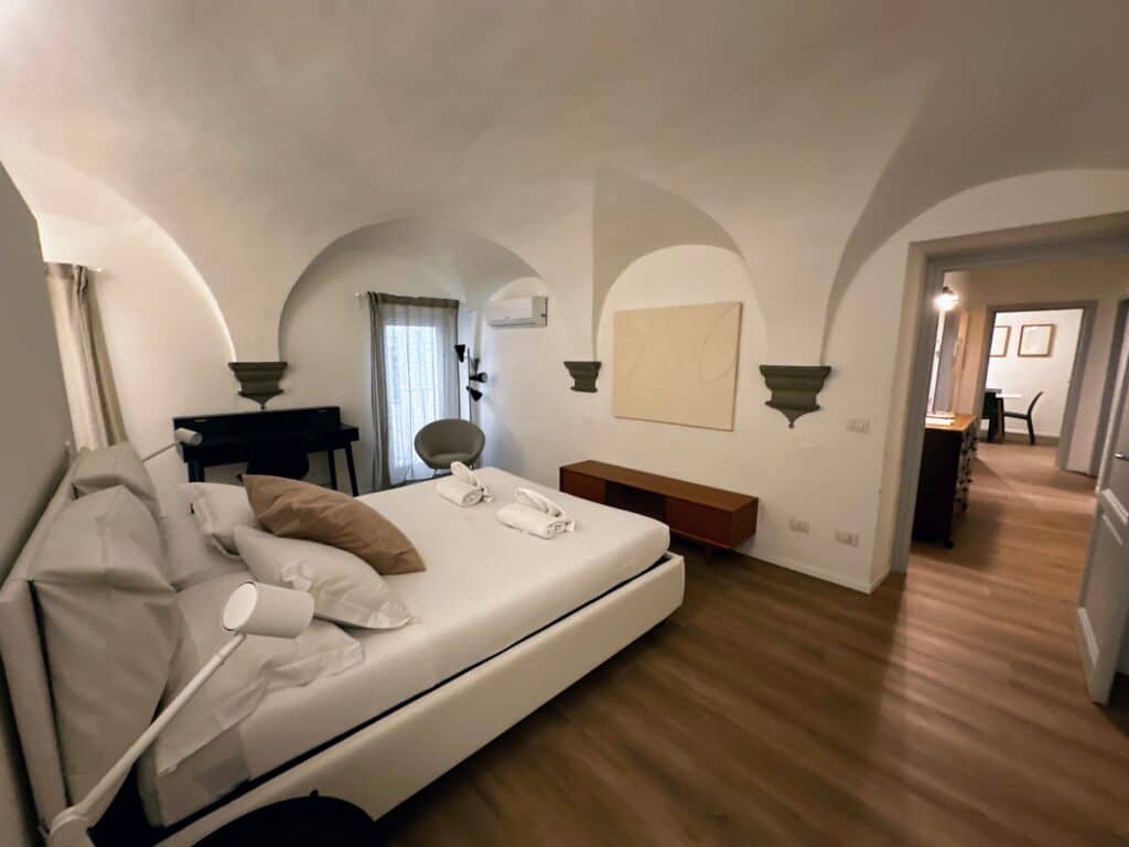 Bedroom with white bed, white walls, and arches in ceiling in an apartment in Florence, Italy. Doorway on right leads to hallway.