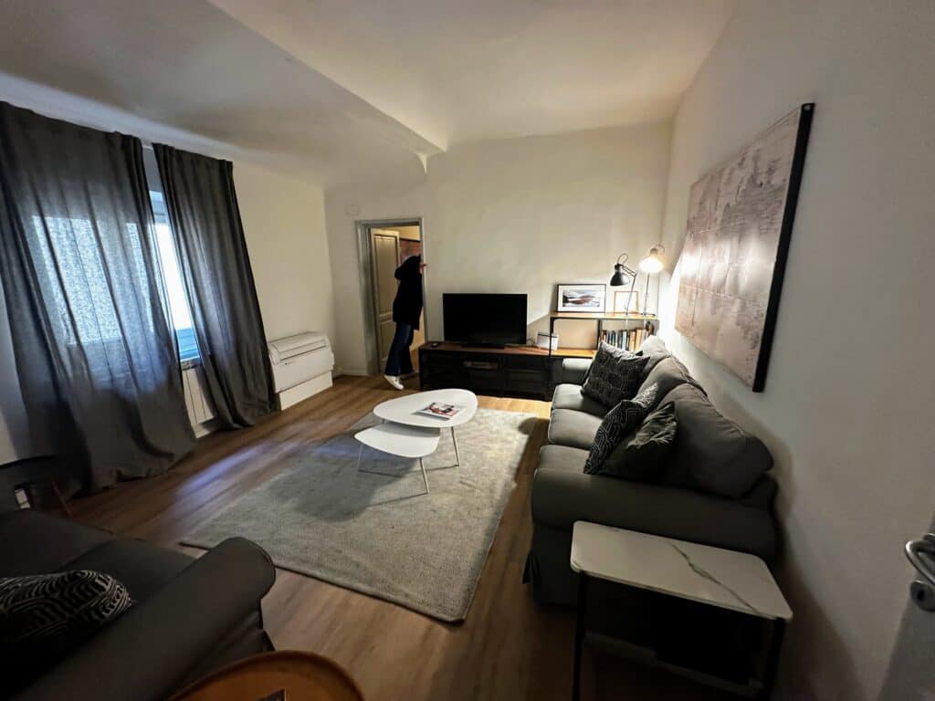 Living room in an apartment in Italy. Couch on right, tv and wooden stand in back. Draped windows on left and woman looks through back door to another room.