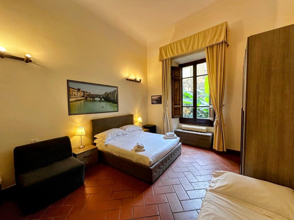 Tile floor, bed, sofa bed, in a room in Florence, Italy. Window looks out onto courtyard and green palm. Painting of Ponte Vecchio behind the bed.