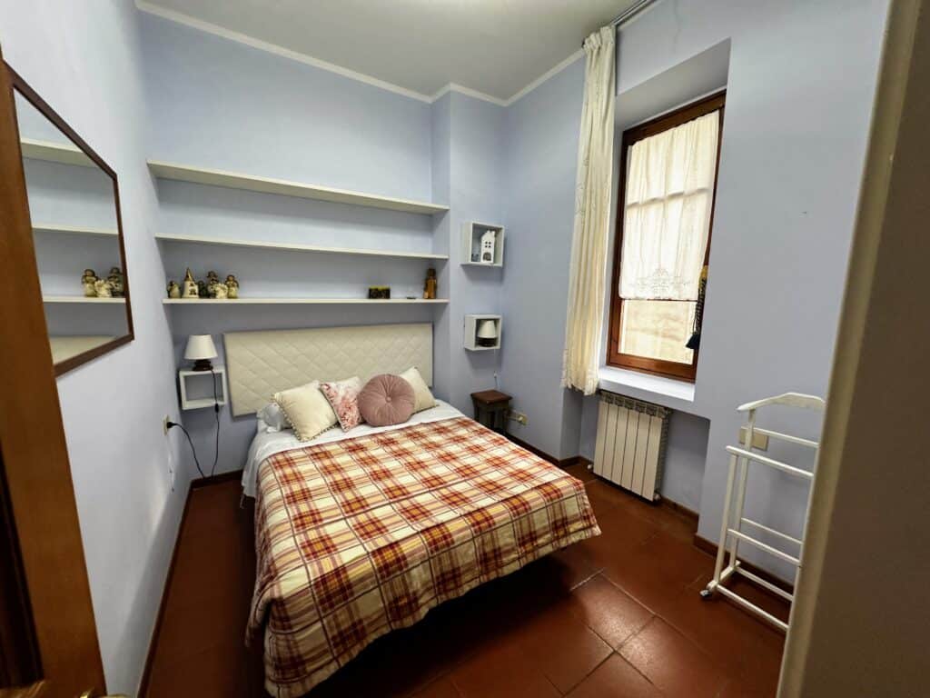 Bedroom in an apartment in Florence. Bed with plaid bedspread and shelves behind it. Window on right, mirror on left. Terracotta flooring.