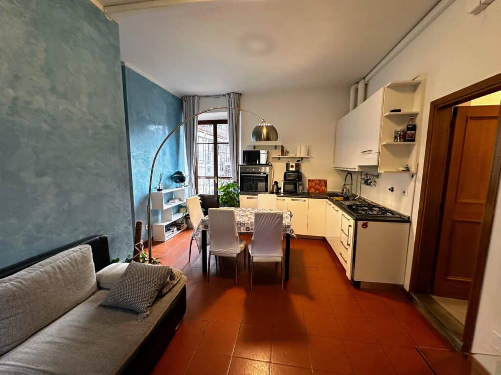 Living area and kitchen in an apartment in Florence, Italy. Blue wall on left, sofa on left, and kitchen ahead. Door open on right.