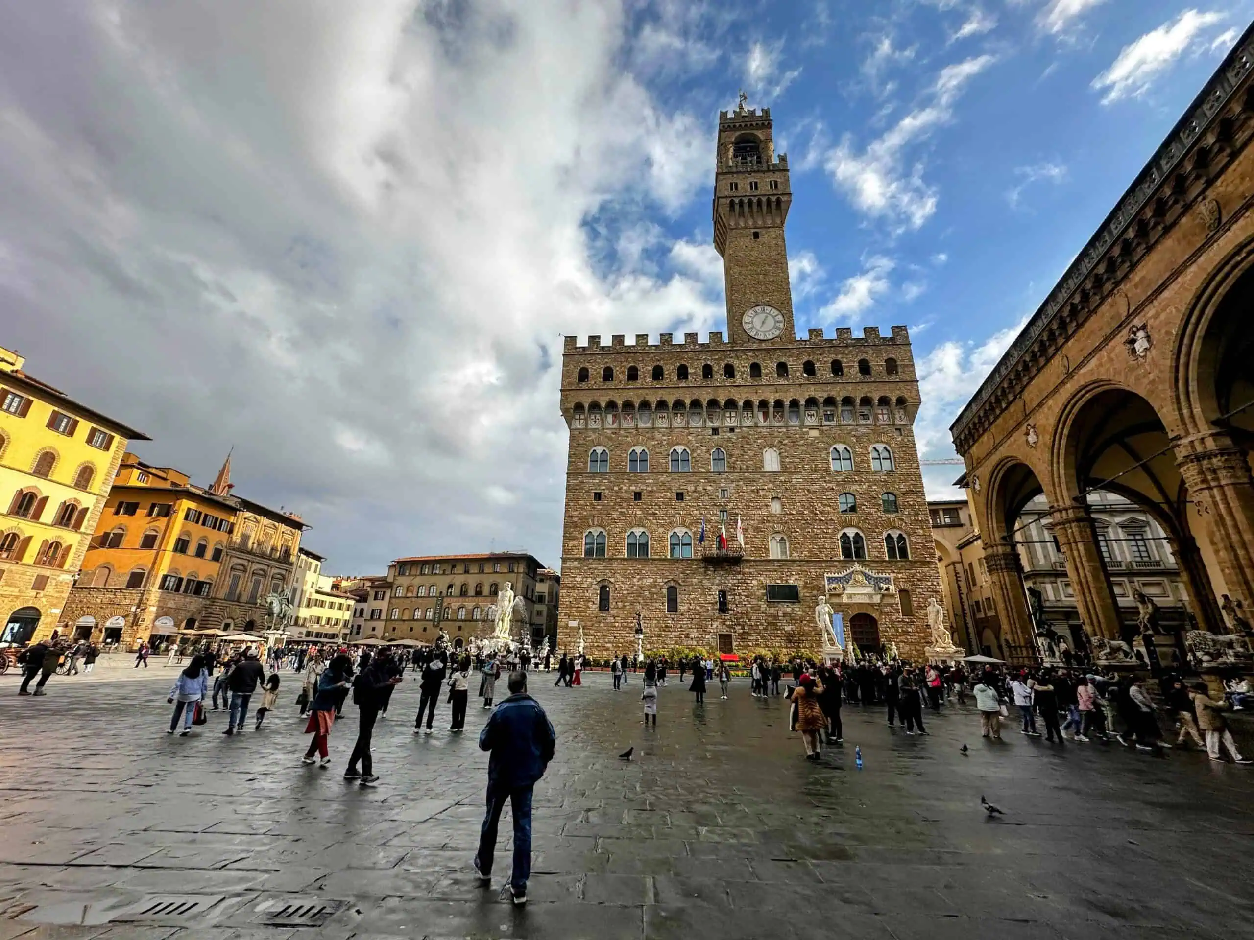 Sunny day with white clouds in Florence, Italy's Piazza della Signoria. Palazzo Vecchio and its Arnolfo Tower are in the center. People walk around the piazza in coats.