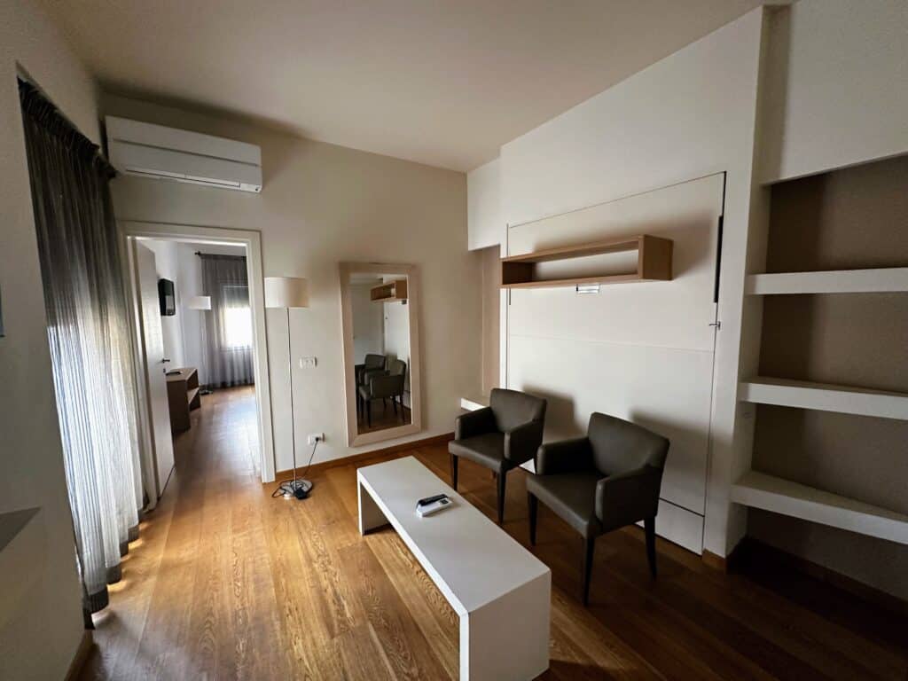 Living area in apartment in Florence, Italy. Two brown chairs with a white table. Mirror in back and door leading to bedroom. Shades on left covering large window.