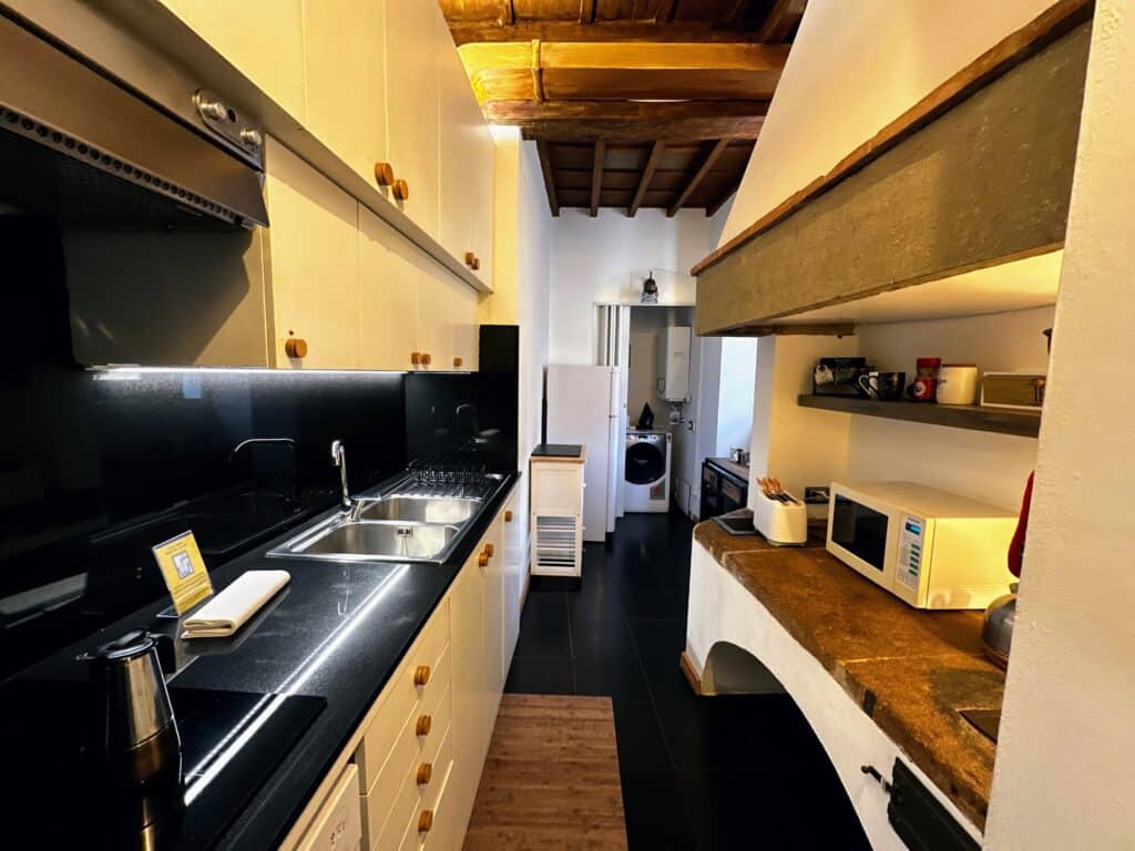 Narrow kitchen in Florence, Italy apartment. Black counters on left with sink and stove and stone counter on right with microwave. Door in back leads to laundry room.
