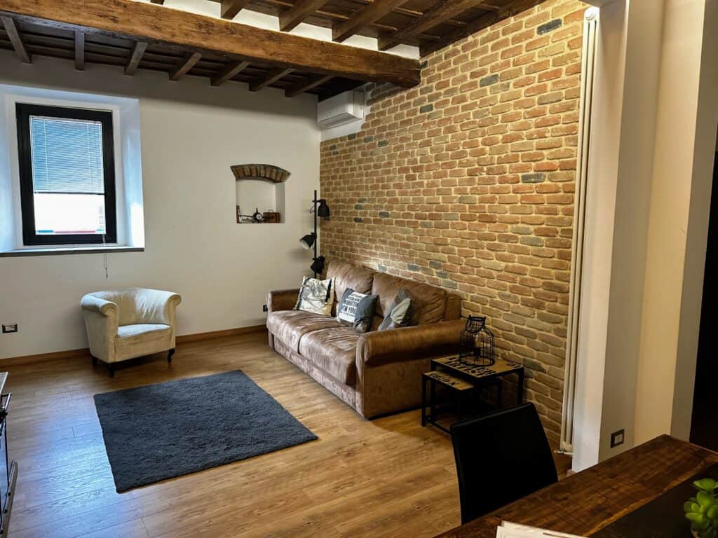 Living room in apartment in Florence, Italy with brown couch, exposed brick wall, and window in back left.