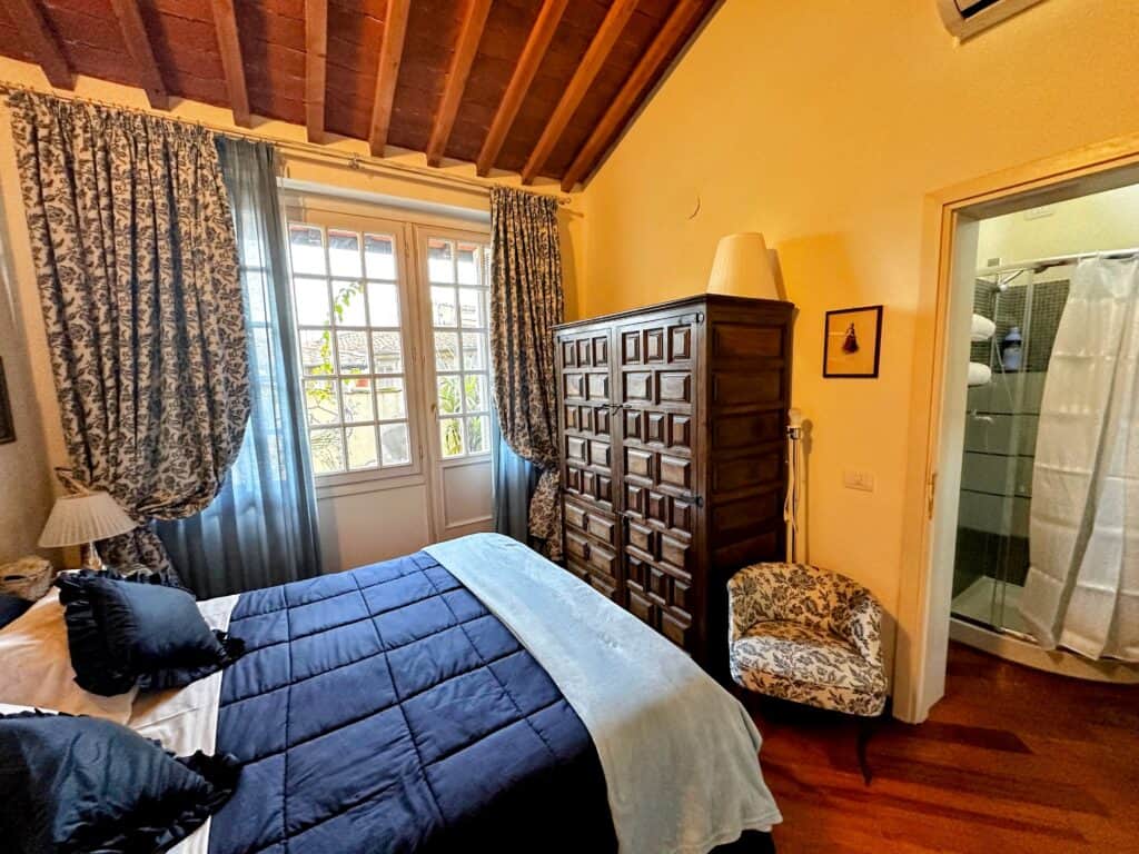 Bedroom in a Florence, Italy apartment with blue bed, wooden dresser, large windows with shades, and door leading into bathroom.