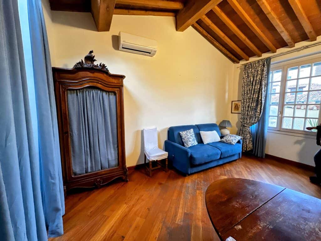 Florence, Italy apartment living area with blue sofa bed and wooden furniture. Wooden beams on ceiling. Window on right looks out on a rooftop.