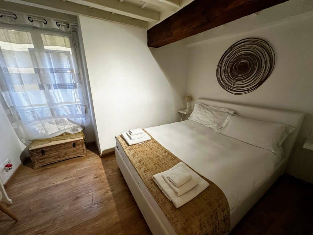 Bedroom in apartment in Florence, Italy. White bed with towels on end of bed. Circular art above bed. Window on left with drapes.