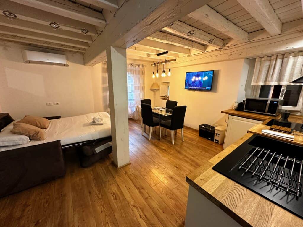 Bright apartment living and kitchen area in Florence, Italy. White painted wooden beams and ceiling. Sofa bed open on left. Dining room table and tv to the right. Window in the back.