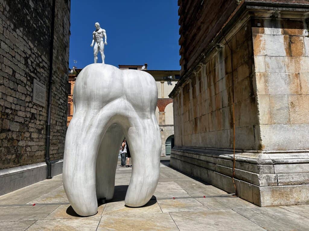 Gigantic tooth statue with a muscular man statue on top of it next to the bell tower in Pietrasanta, Tuscany.