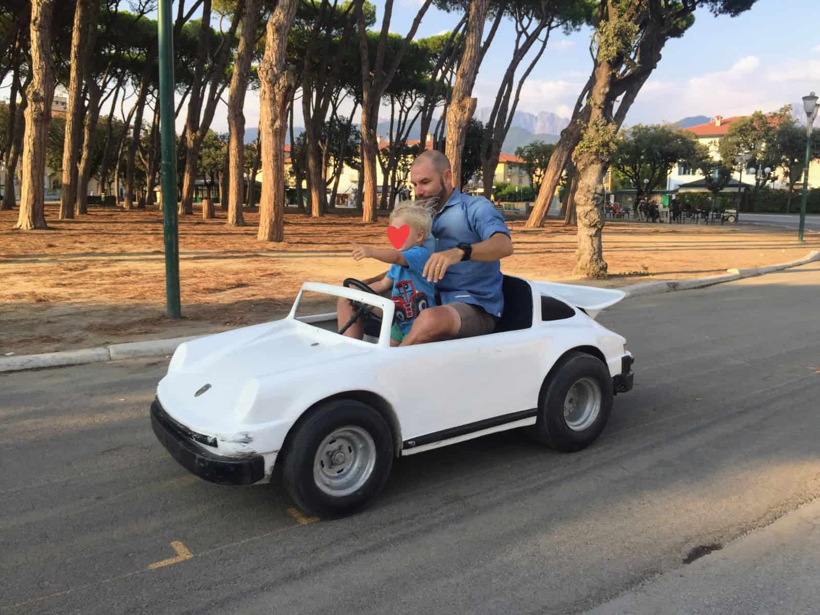 Father and son ride in a small white car next to an umbrella pine forest in Forte dei Marmi, Italy.