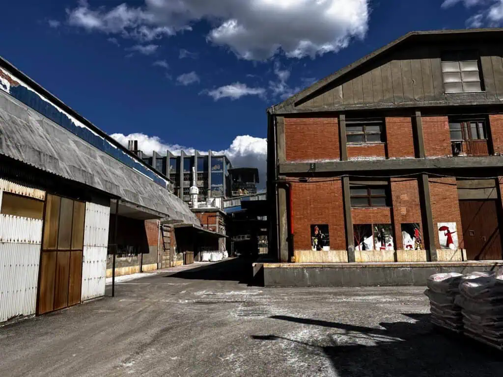 The outdoor area of the Saline di Volterra. Large buildings and paved roads with sprinklings of salt. Sunny day with white clouds. Building on right has photos of exhibitions and performances inside the salt factory.