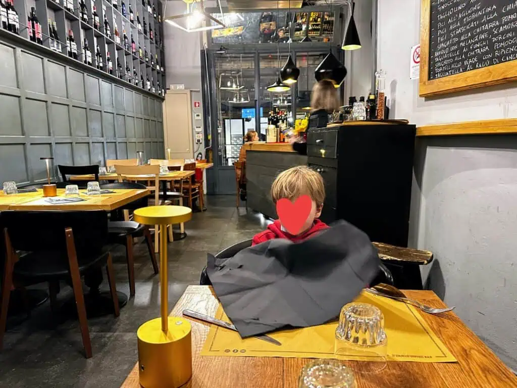 Boy sits at wooden table and opens a black napkin onto the table at a restaurant in Florence, Italy. There are other tables but no one is sitting at them. Chalkboard with menu on right and bar area behind the boy.