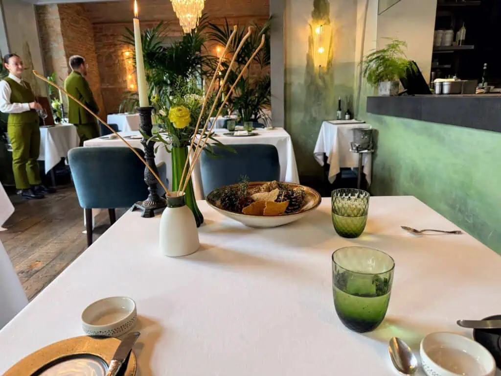 Dining room table at Saporium restaurant in Florence. Waiter and waitress stand in background. Opening window of kitchen on right. Table has bread bowl, breadsticks, green glasses.