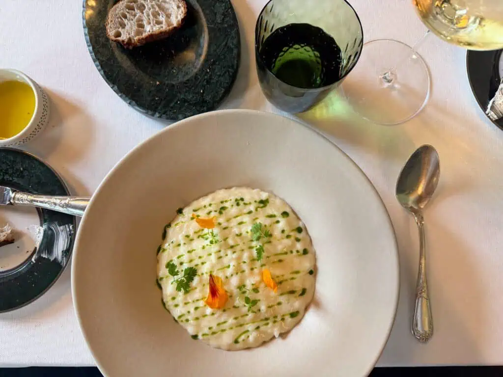 White tablecloth with white dish of risotto. The risotto has green droplets in a linear pattern. Also green herbs and yellow petals. Around the plate of risotto are silverware, bread dishes, water glass, and wine glass with white wine.