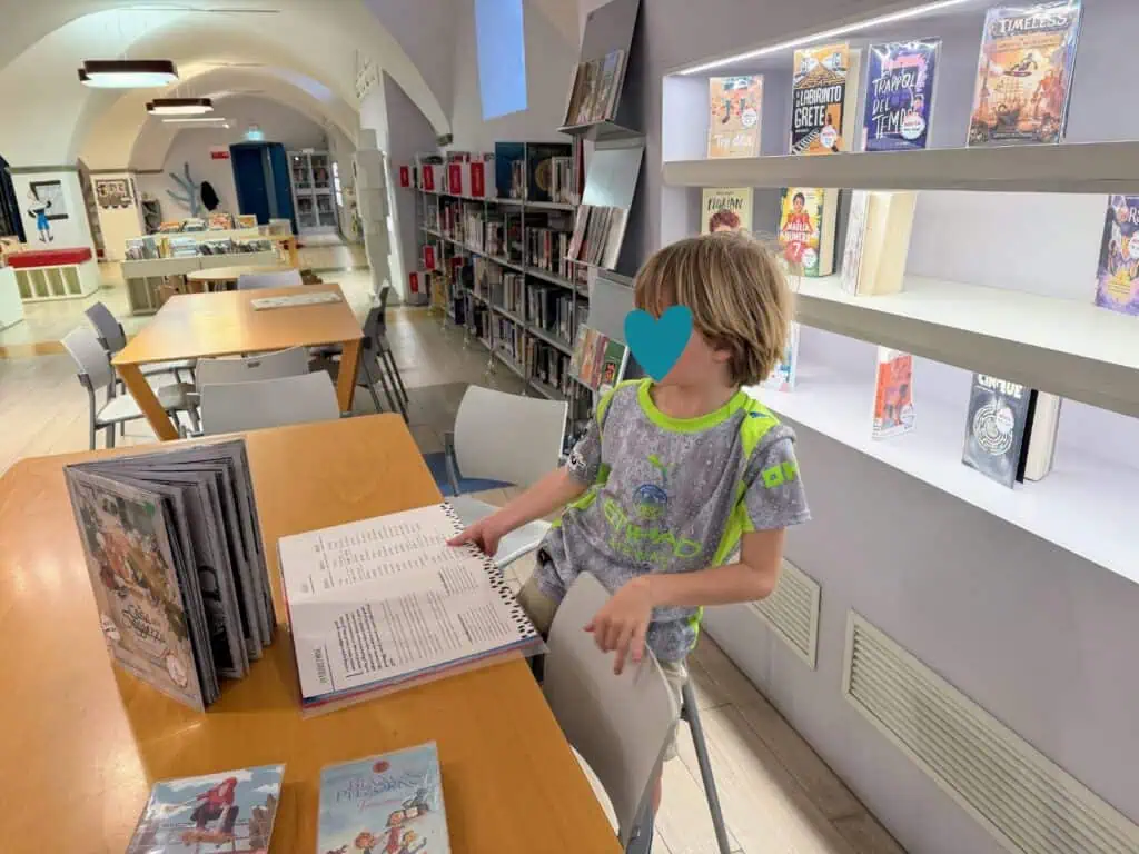 Boy looks at open book while he stands at a table inside a library. He's in the children's section and there are chapter books on the shelves behind him.