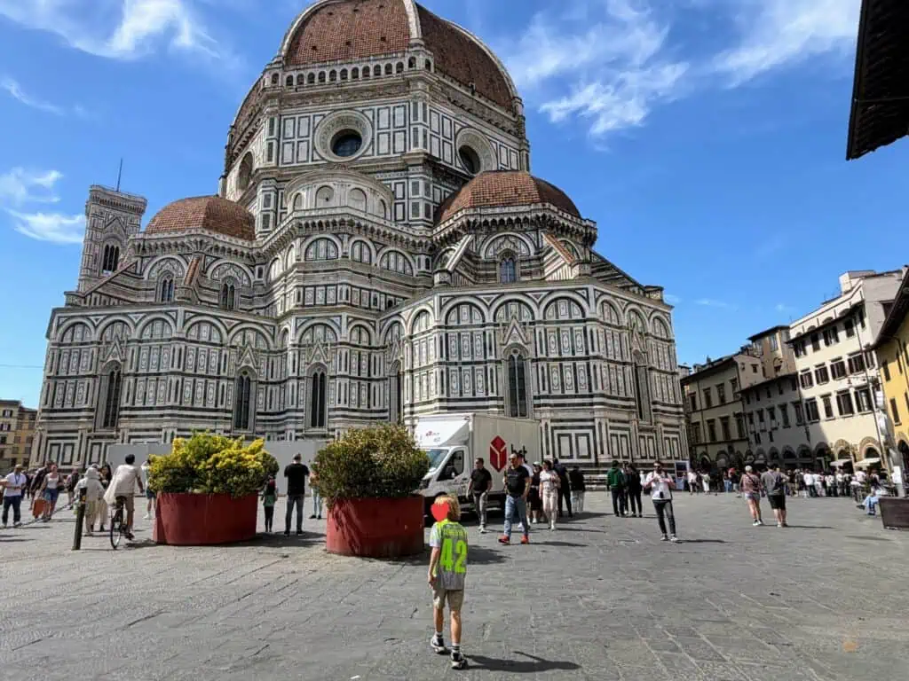 Boy walks in front of the Duomo in Florence, Italy. Other people are also walking near it. It's a sunny day with whispy white clouds.
