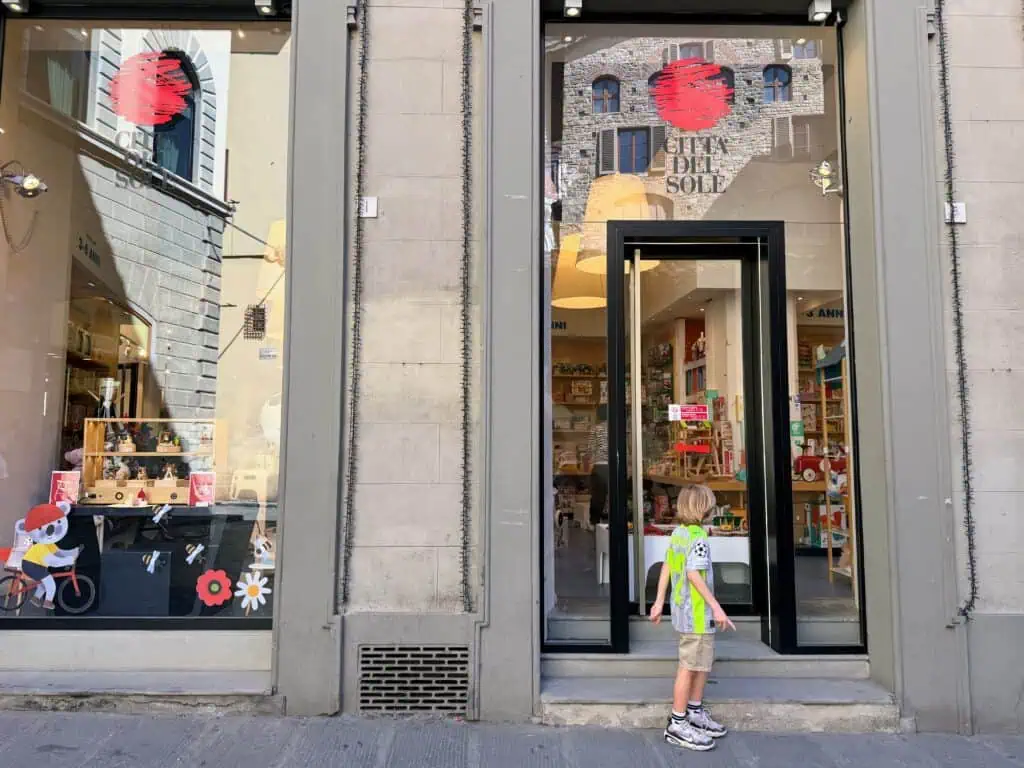Boy stands in front of glass doorway to toy shop in Florence, Italy and he looks inside at the toys. The logo of the shop, Città del Sole, is painted on the upper glass windows.