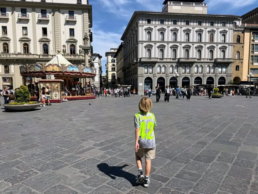 Boy in Semenyo soccer jersey walks in the middle of Piazza della Repubblica in Florence. Historic carousel ahead of him on left and multi-story buildings ahead of him. Others walk in the far end of the piazza.