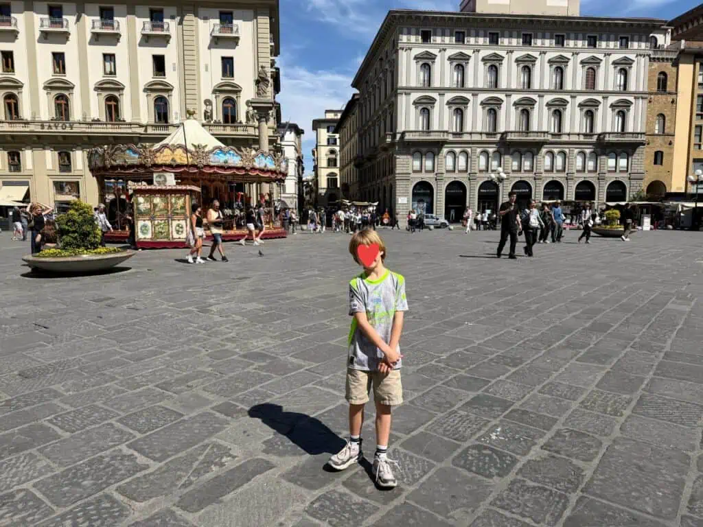 Boy stands in the middle of Piazza della Repubblica in Florence, Italy. Behind him are multi-story buildings lining the piazza and a historic carousel. Some people are walking at the far end of the piazza.