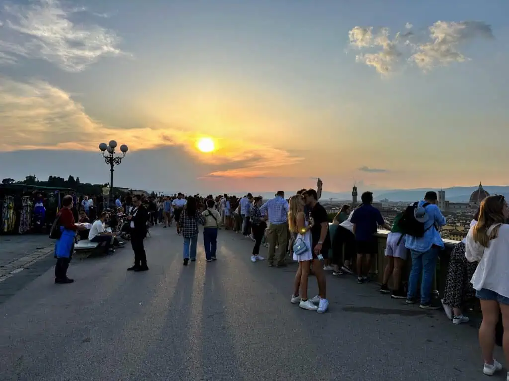People standing in a paved area looking at the sunset over Florence, Italy. Vendors on left.
