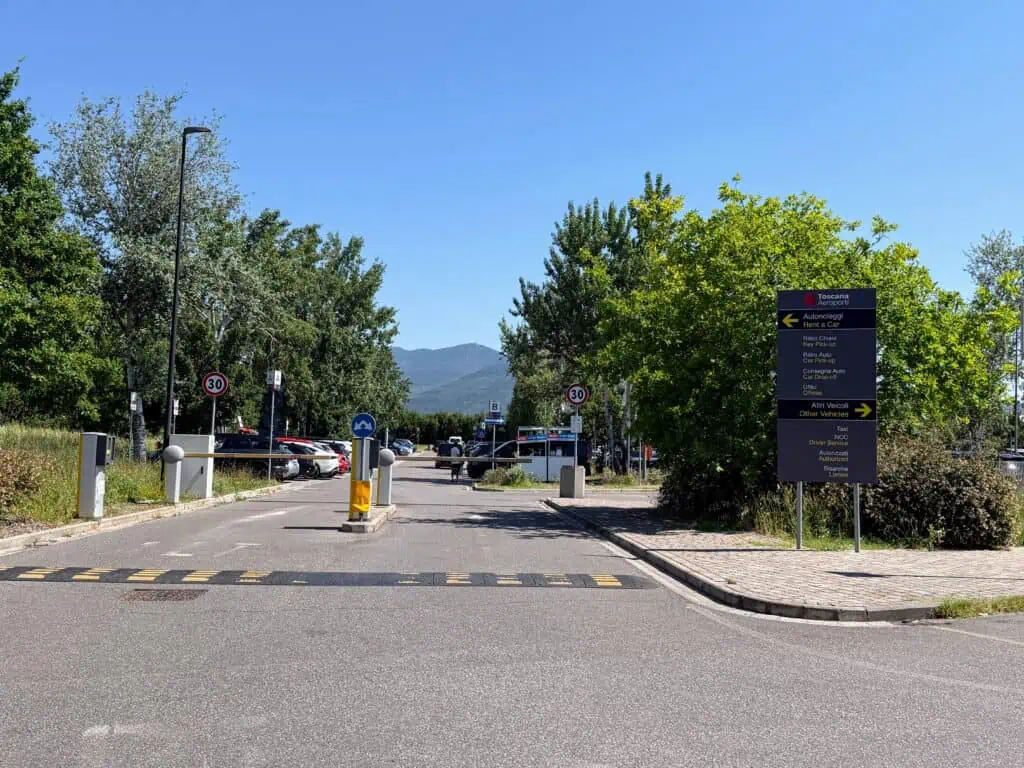 Entrance to parking lot with speed bump and barriers that can be raised. Grey sign on right. Parked cars in lot behind the barriers. Trees and bushes around the lot.