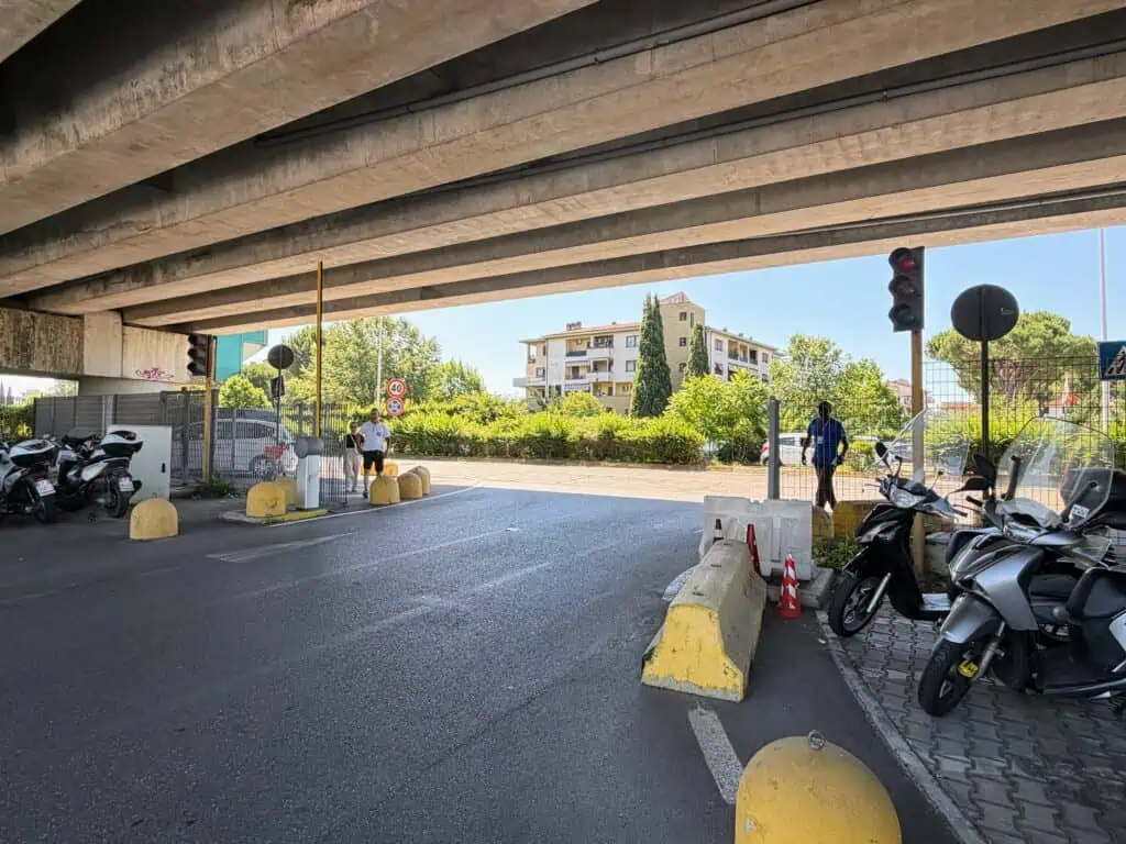 Paved road leading to a T-intersection. The road is covered by an overpass. There are motorcycles parked on the right and left. A few people stand by the road. There is a traffic light but it's not lit. This is at the Florence, Italy airport car rental lot.
