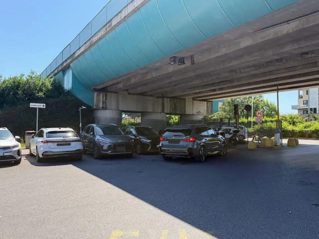 Cars are parked under and overpass. There is a road to the right. A white sign for uscita-exit is on the left. This is at the Florence, Italy airport car rental lot.