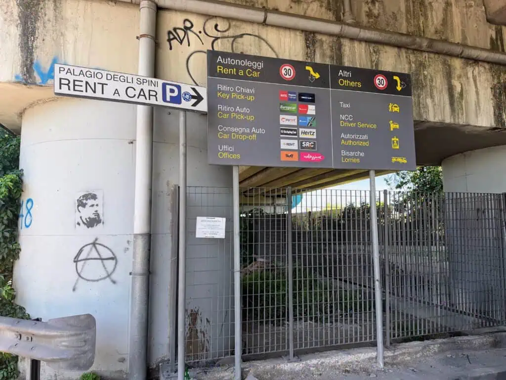 Grey signs for car rental location at the Florence, Italy airport. They're attached to the bottom of an overpass. There's graffiti on the wall and also a sign for 'Palagio degli Spini - RENT a CAR' to the left.