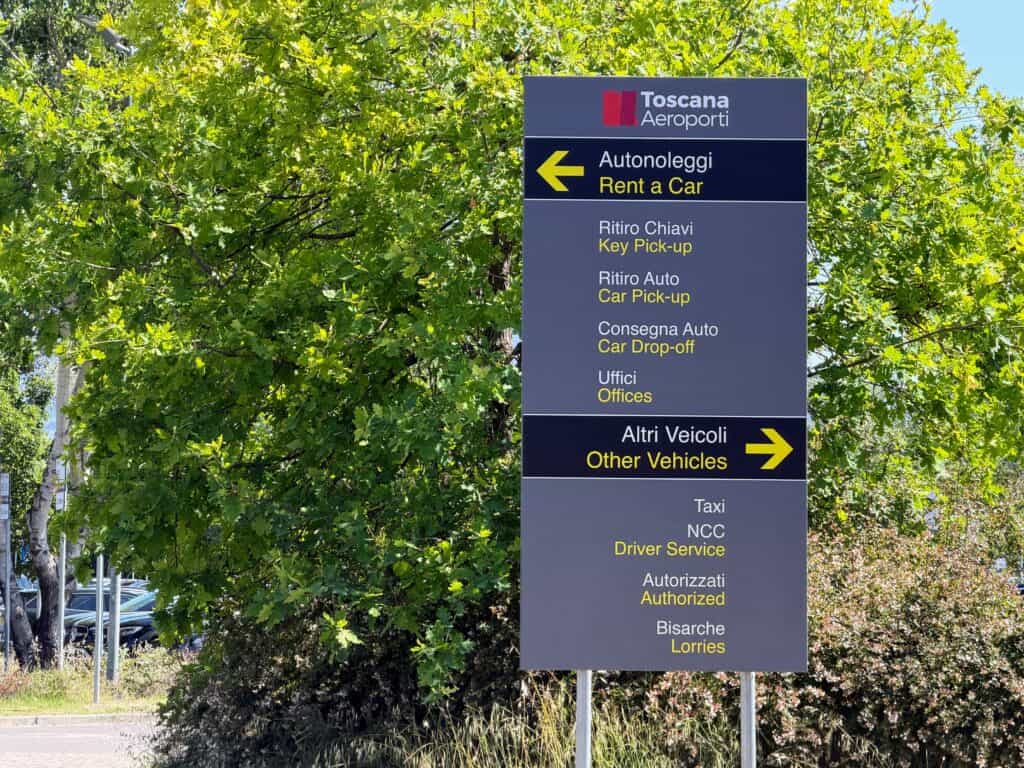 Large grey sign in front of leafy tree at the Florence, Italy airport car rental locations. Arrows point to where drivers should go.